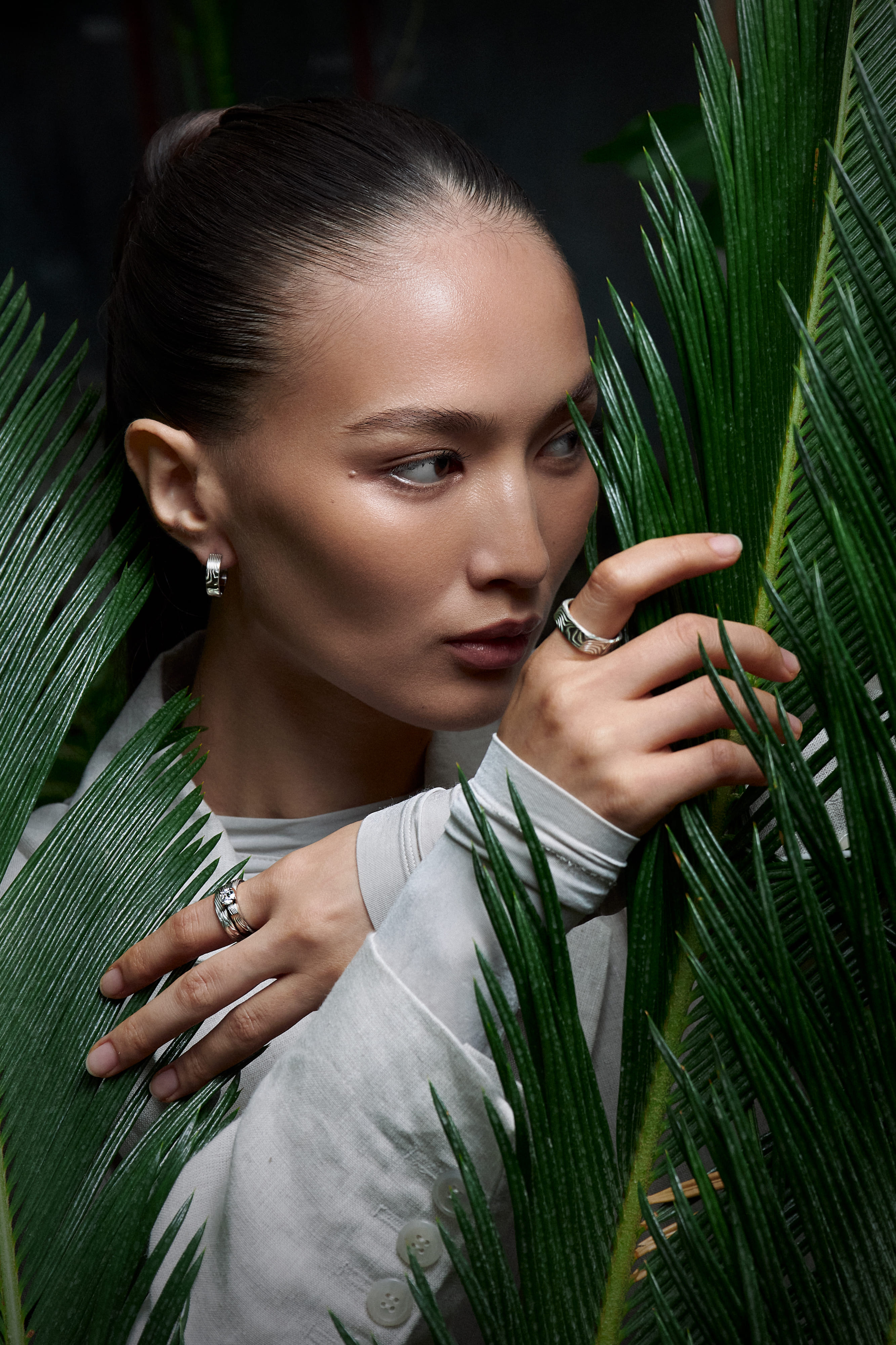 Woman among green leaves wearing jewelry from Kochut’s The Bloom of Time collection: Yutori earrings, Tenno ring, Kanso wedding bands, and Empress Kojun ring crafted from palladium gold, white gold, and etched silver.