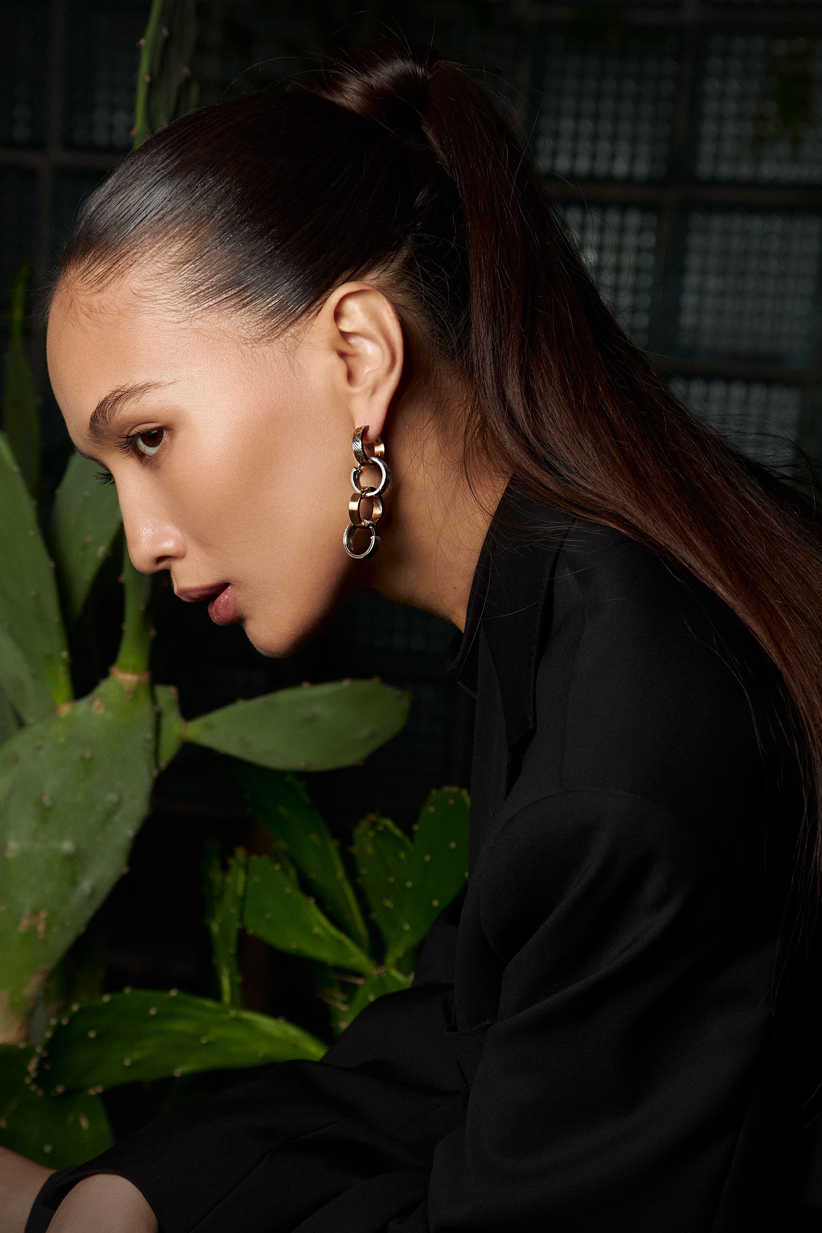 Woman in a black blazer wearing Yutori earrings from Kochut’s The Bloom of Time collection, mokume earrings in red gold with etched and oxidized silver, modern portrait with cactus background.