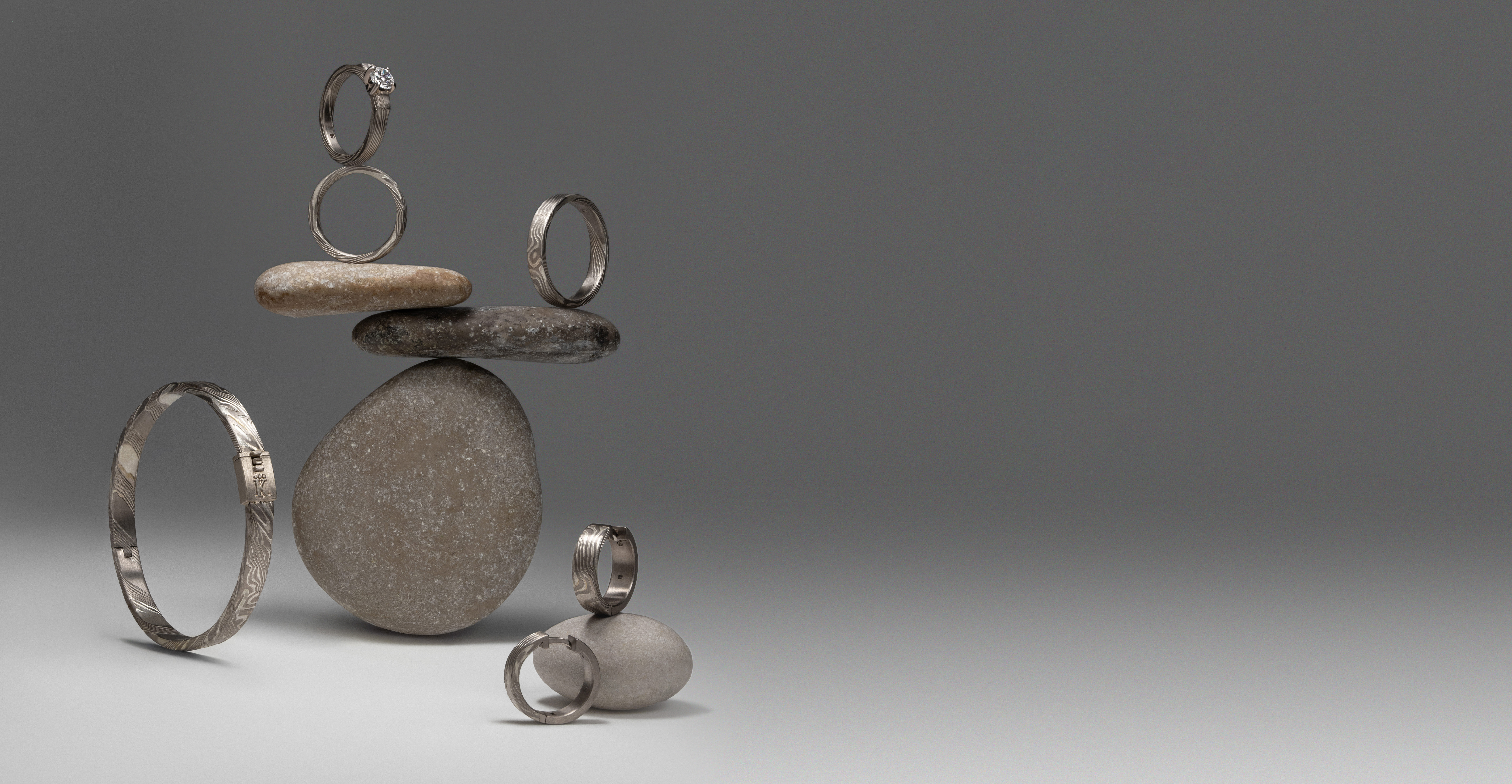 Jewelry from “The Bloom of Time” collection in Mokume-gane technique: Enso bracelet, Tenno ring, Empress Kojun ring, and Kado and Kanso wedding bands, displayed on stones in a minimalist composition.